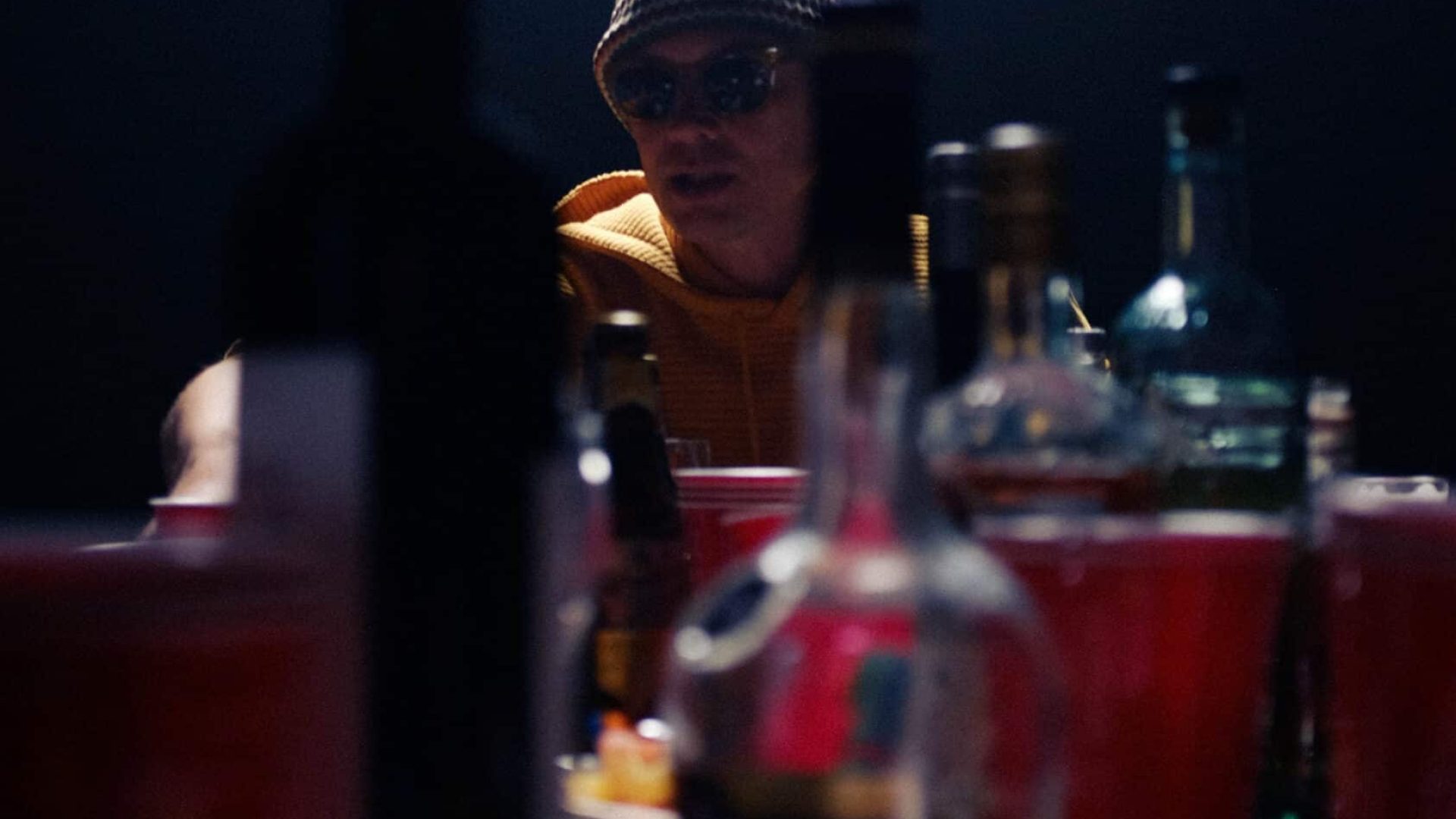 Person wearing a hat and sunglasses at a table with bottles and food, dim lighting.