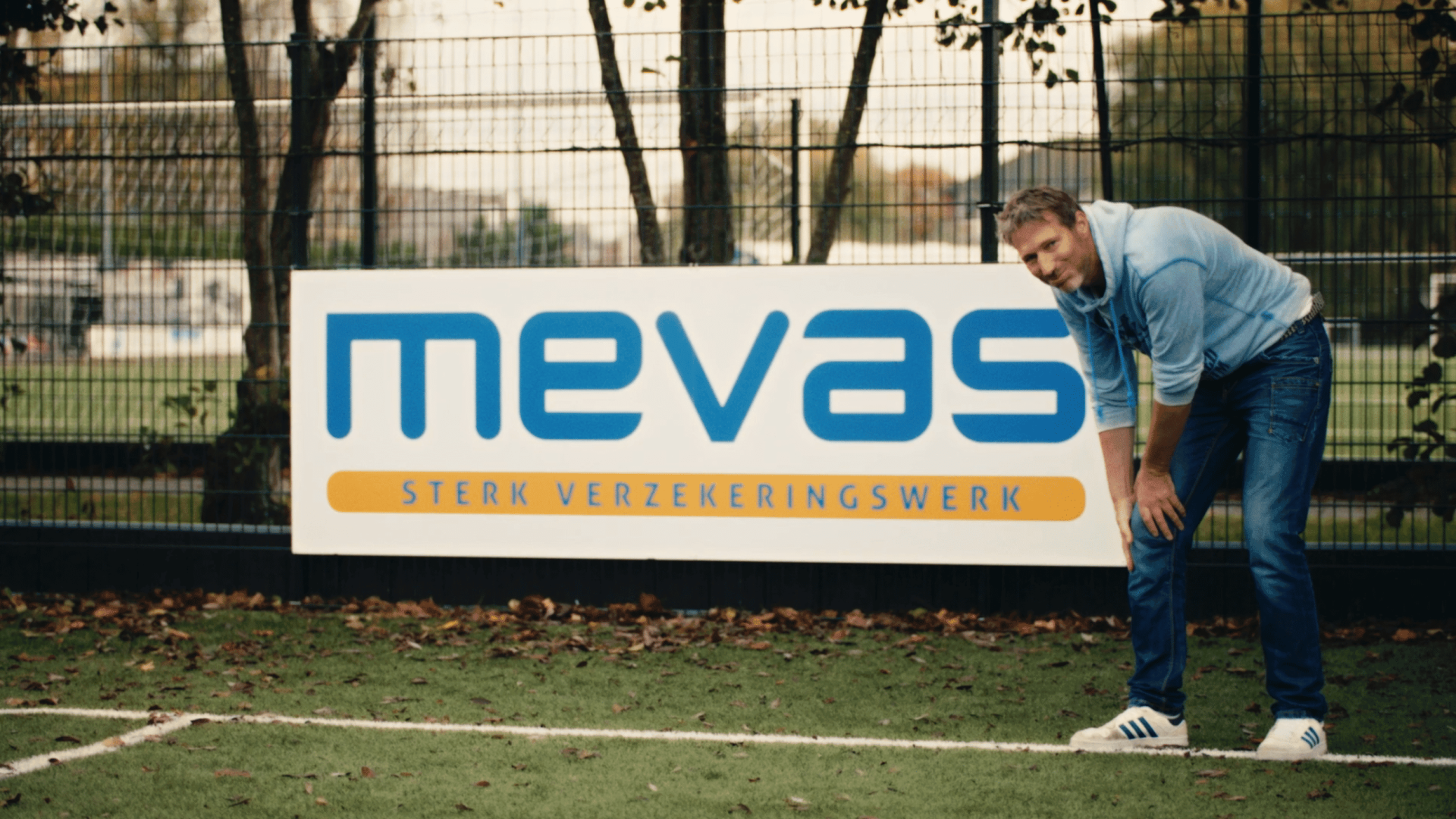 Man measuring a tennis court with a "Mevas" insurance sign in the background.