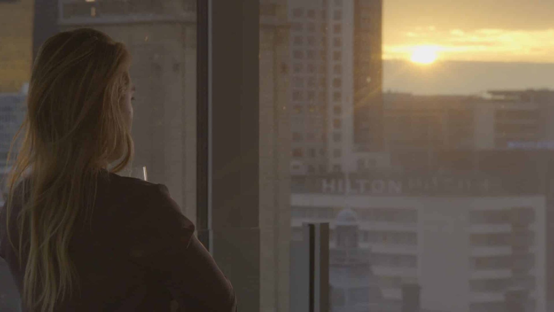 Woman looking out window at sunset in city skyline, creative video agency scene.