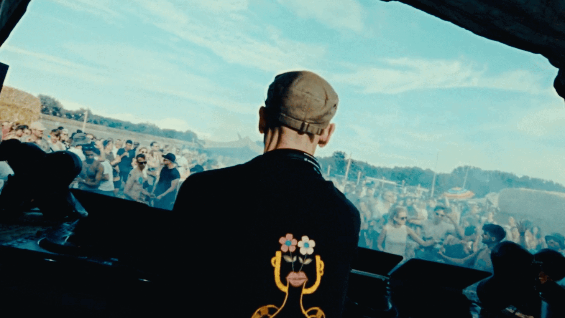 DJ performing at outdoor music festival with a lively crowd and scenic sky backdrop.