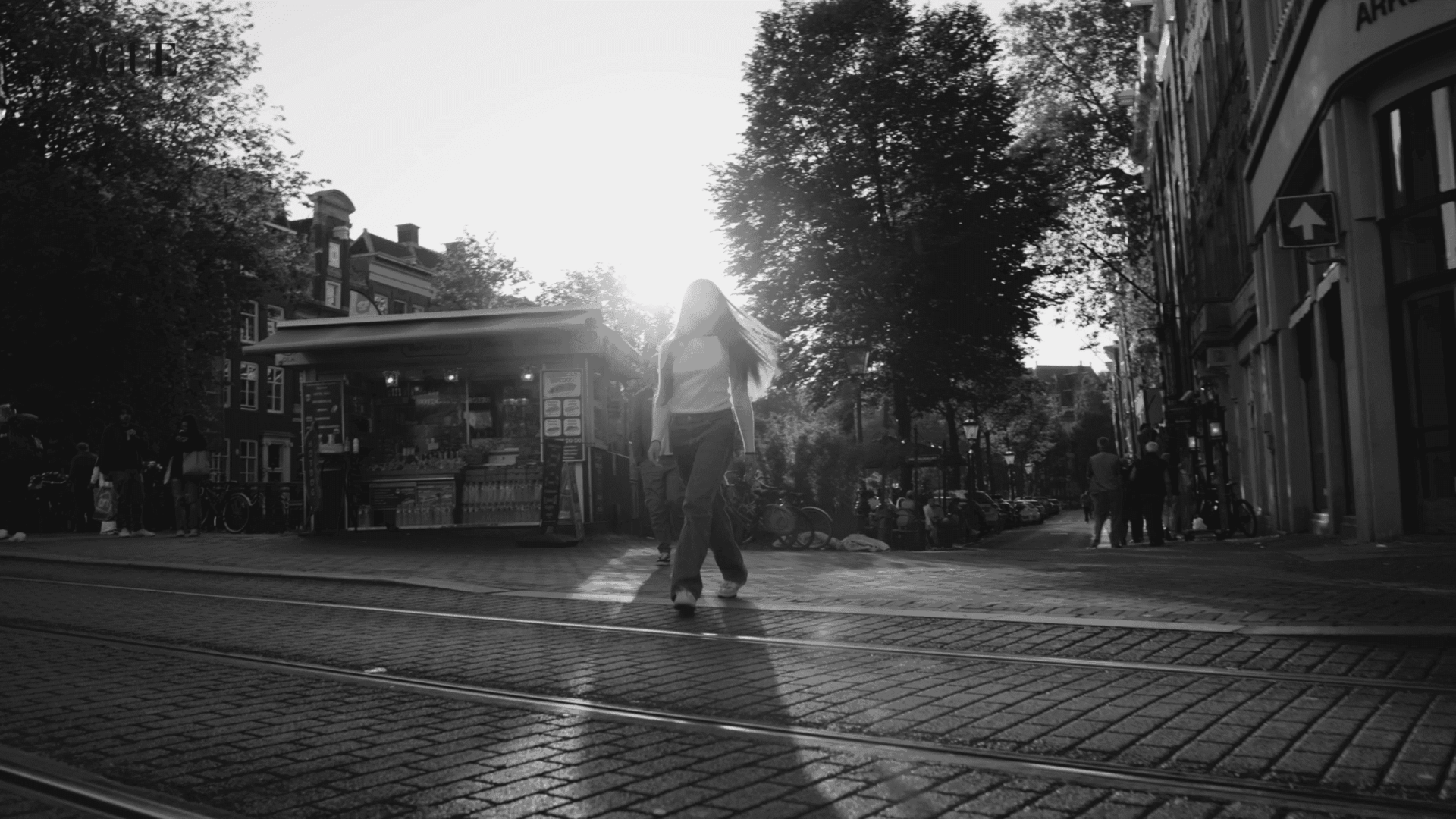 Woman walking on city street at sunset, urban scene with tram tracks and trees.