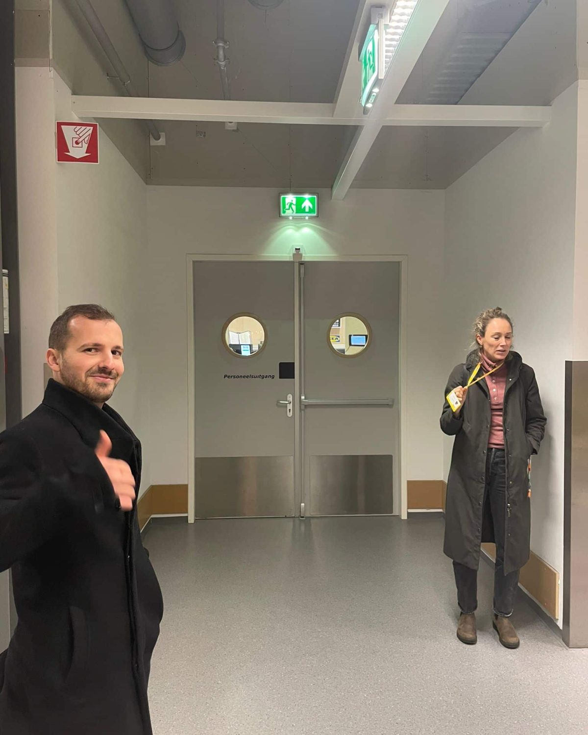 Two people standing in a hallway, one giving a thumbs-up, the other holding a drink.